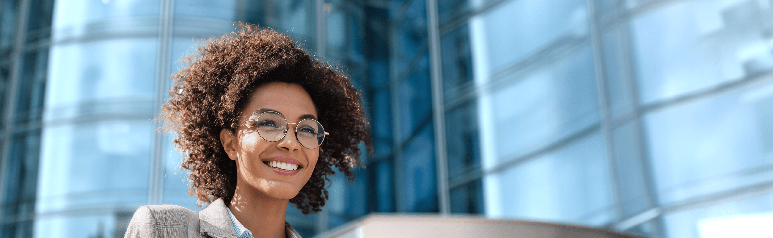 Professional with curly hair and glasses smiling while holding a mobile device against modern office building backdrop